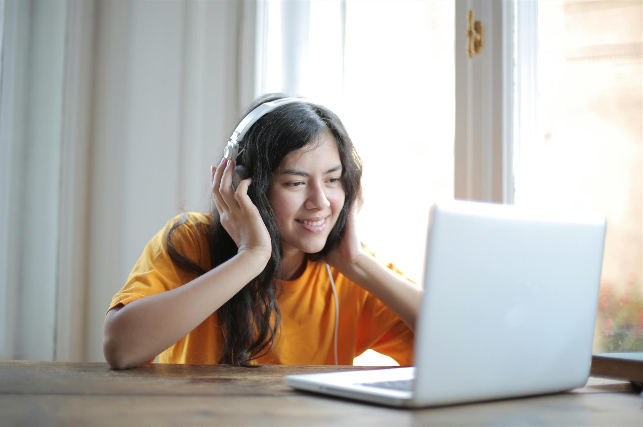 A person wearing headphones smiles while looking at a laptop. They are seated at a wooden table near a bright window, suggesting a relaxed, indoor setting.