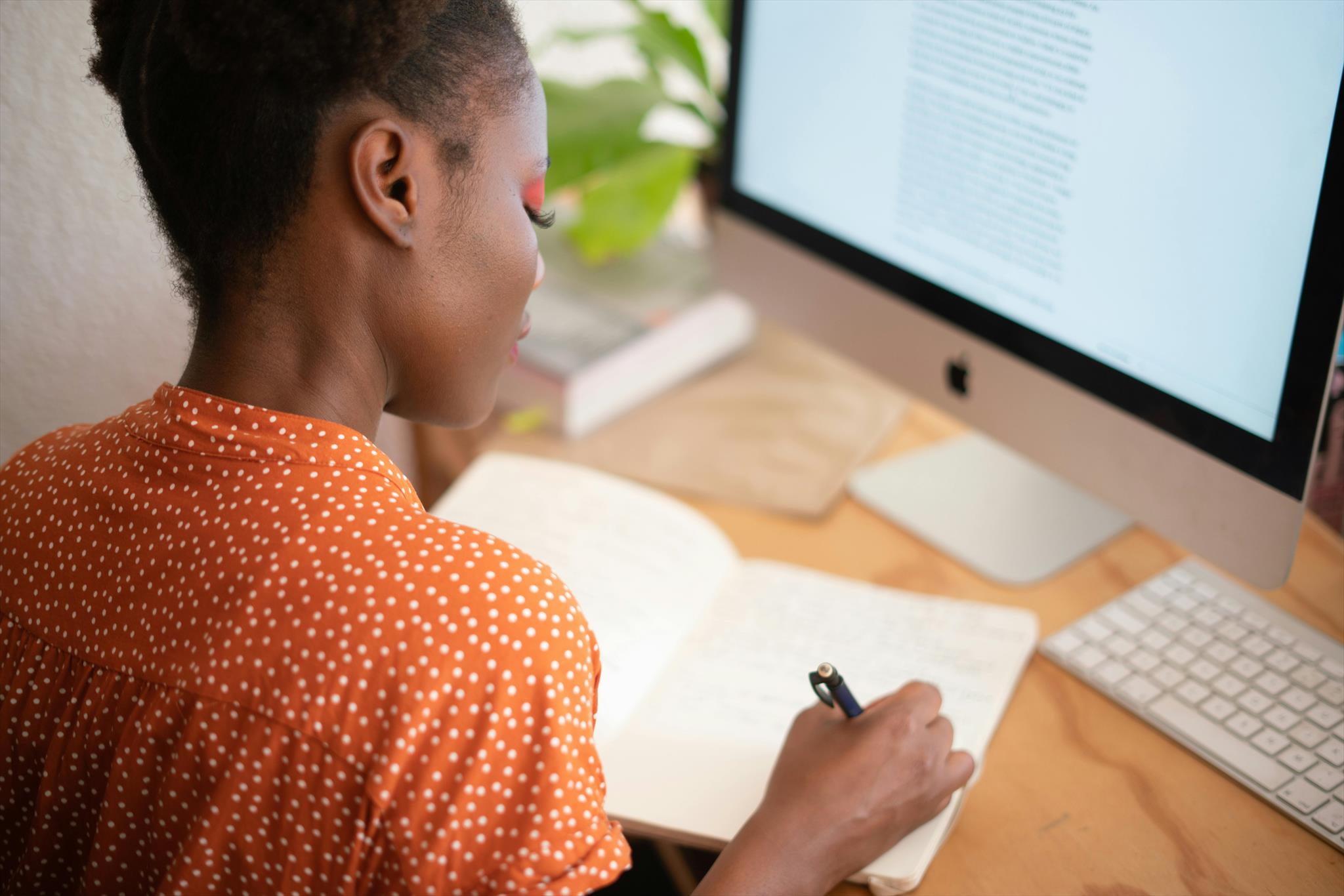 A person in an orange polka dot shirt writes in a notebook beside a computer displaying text, at a wooden desk with a keyboard.