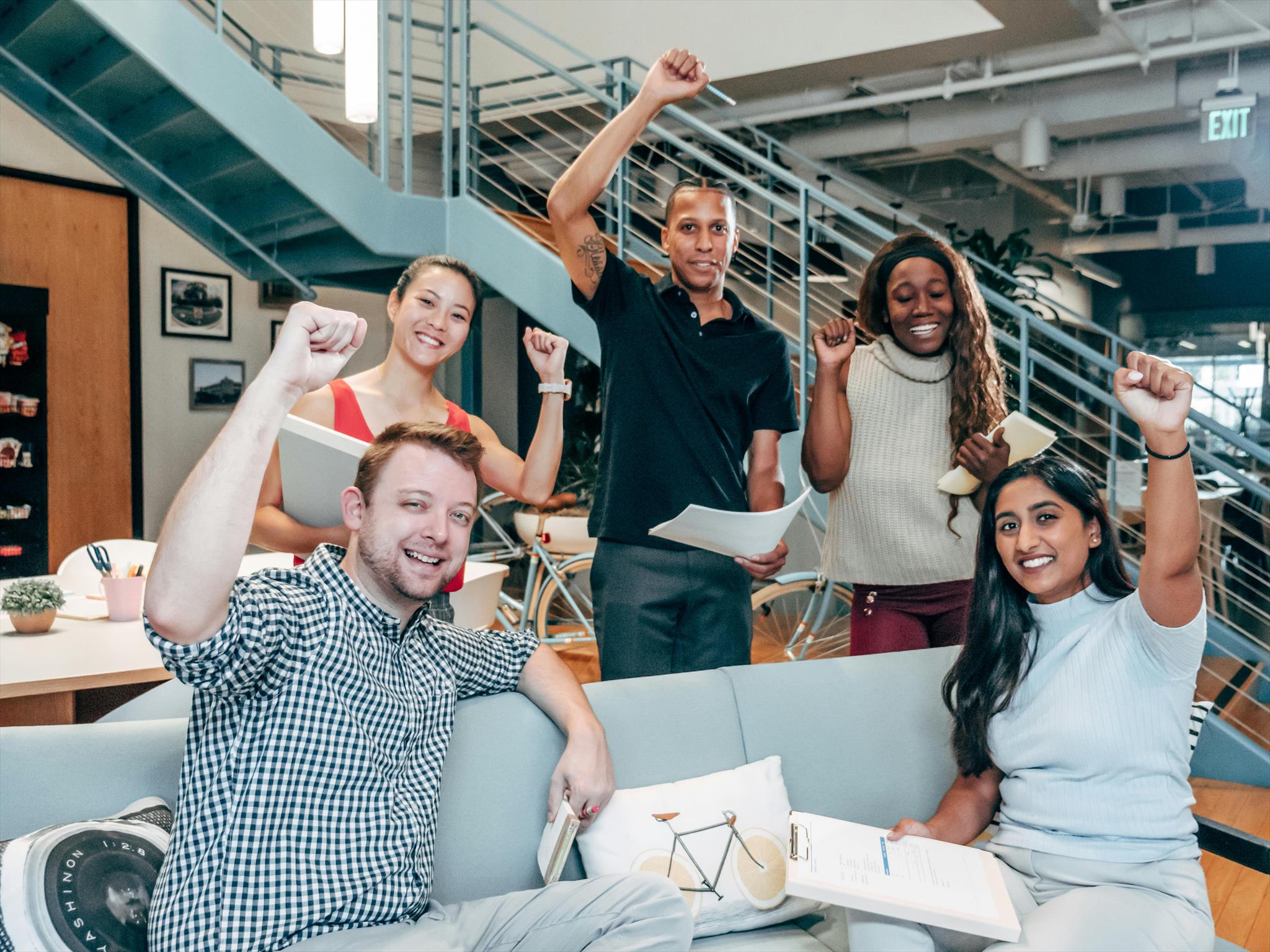 Five people raise their fists in celebration, holding papers, in a modern office with a staircase and framed pictures on the walls. A couch and table with office supplies are nearby.