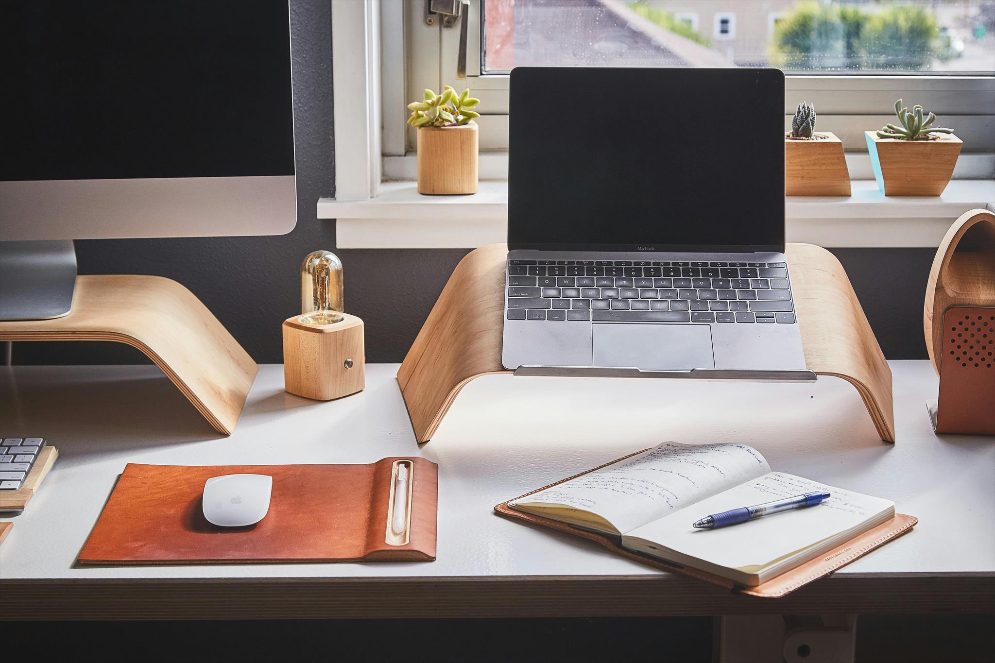 Laptop resting on a curved wooden stand, alongside a desktop computer, notepad, and lamp on a white desk. Succulents in wooden pots by a window in a modern workspace.