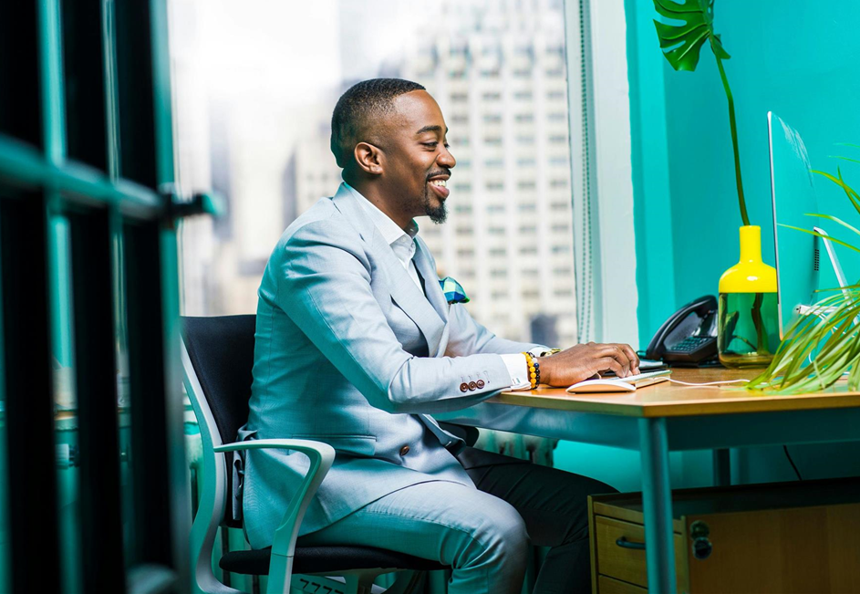 A man in a light suit types on a computer at a wooden desk in a brightly lit office, with a cityscape visible through a large window.