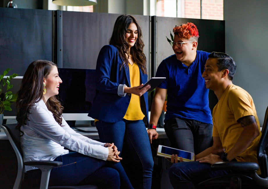 Four people are interacting cheerfully in an office setting. One person is holding a tablet while others are gathered around, smiling and conversing. Computers and plants are in the background.