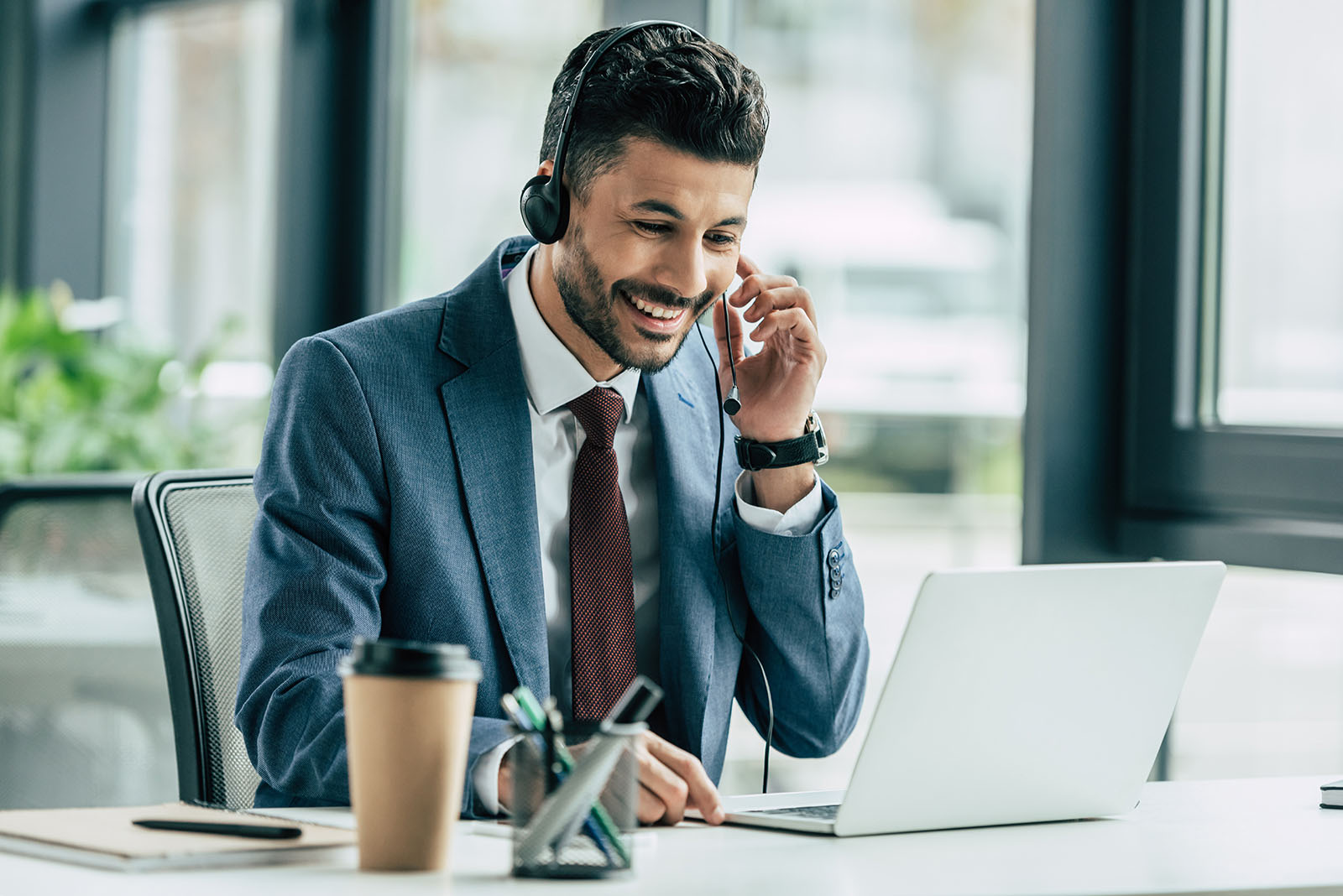 Bonfire Training - Training Solution for Remote Workforce A man in a blue suit talks on a headset while using a laptop. He sits at a desk with office supplies and a coffee cup, in a bright office environment.