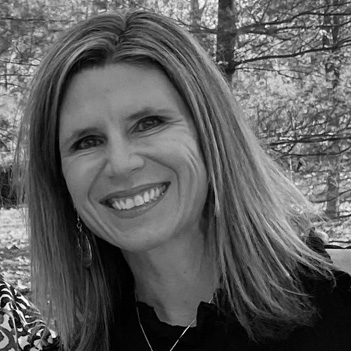 A woman smiles warmly outdoors, surrounded by trees. Her hair is long and straight, and she wears a dark top with an abstract pattern.