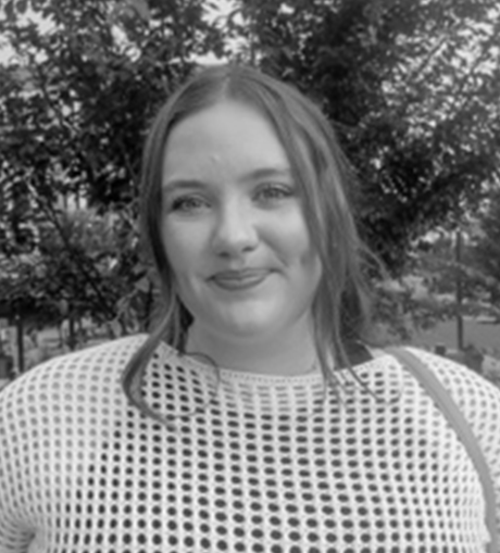 A person smiles slightly, standing outdoors. Trees with leaves form the background, and the person wears a netted top.