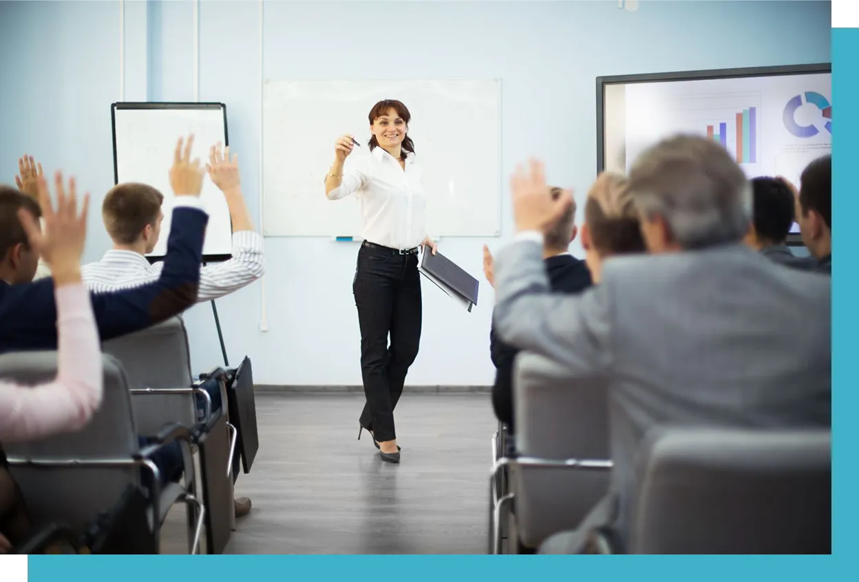 A woman stands in front of a classroom, holding a marker and papers, engaging with an audience, some of whom have raised hands. A whiteboard and presentation screen are visible.