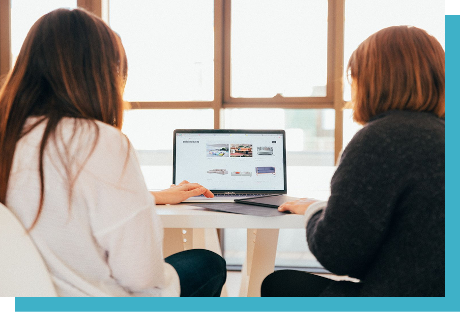 Two people sit at a table, using a laptop displaying a webpage titled "archiproducts," featuring images of home furnishings, in a bright room with large windows.