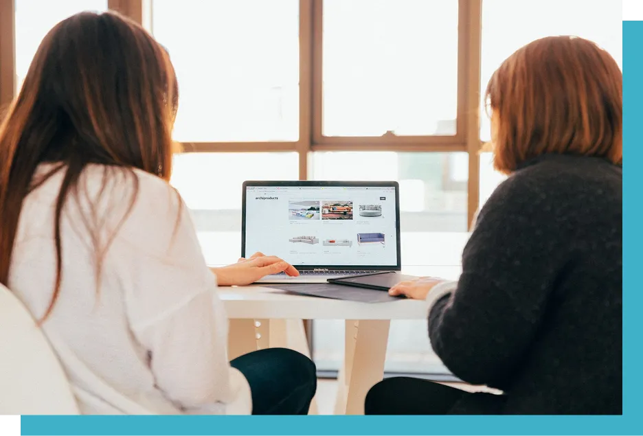 Two people sit at a table, using a laptop displaying a webpage titled "archiproducts," featuring images of home furnishings, in a bright room with large windows.