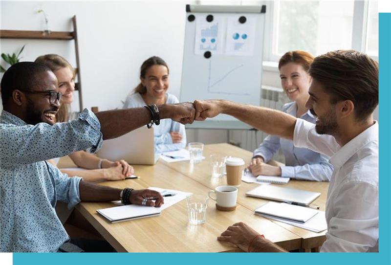 sterch10 People fist-bump across a conference table, surrounded by smiling colleagues with notebooks and laptops, in a bright office with a flip chart displaying graphs.