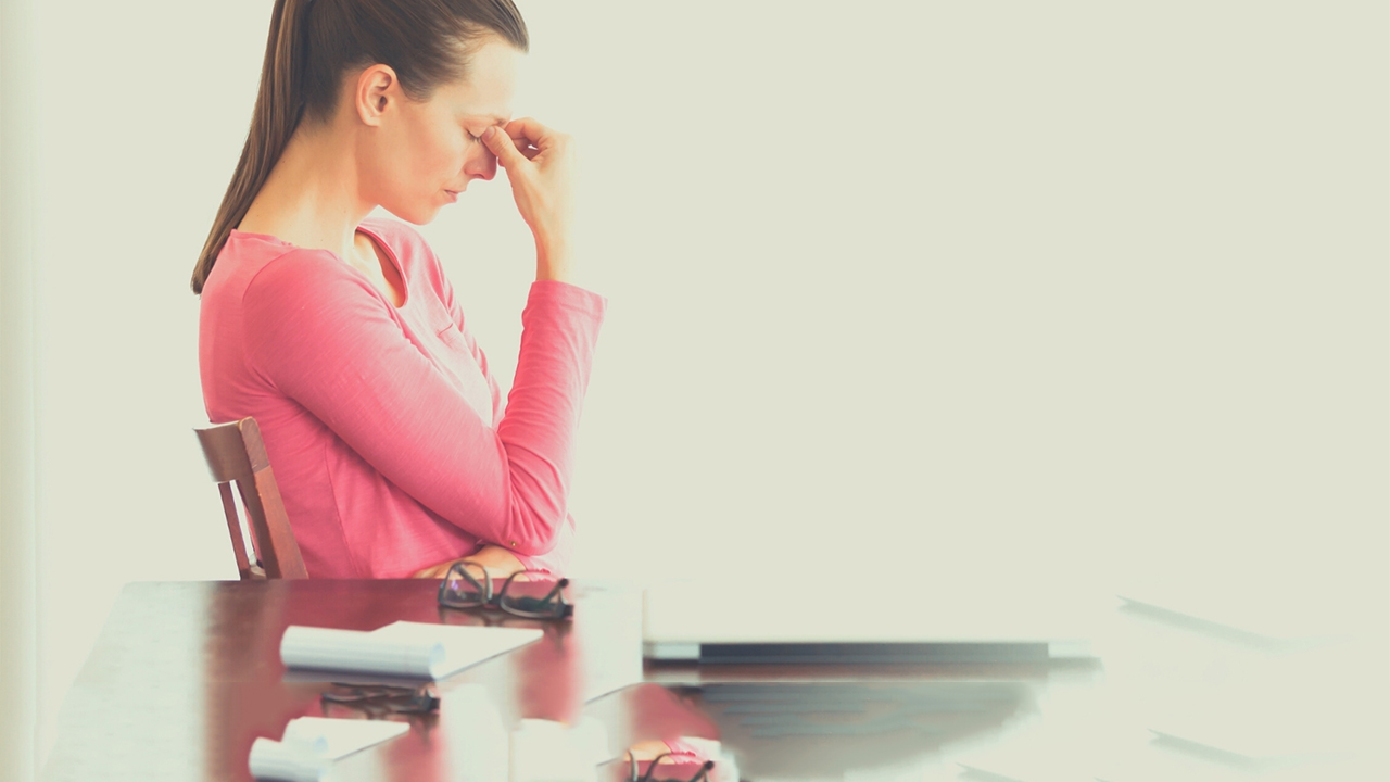 A woman in a pink shirt sits at a table, rubbing her eyes, next to a laptop, glasses, and a notebook, appearing stressed or tired in a bright, minimalist setting.