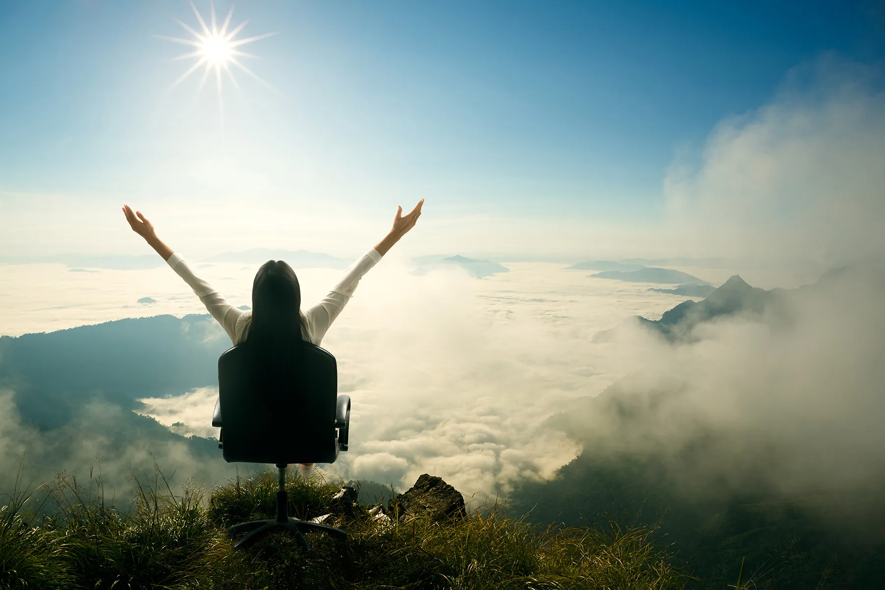 Person sitting in an office chair on a mountain overlook with arms raised, facing the sun above a sea of clouds.