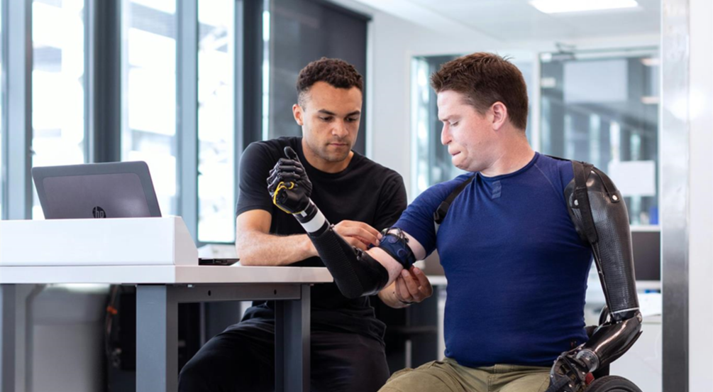A man with prosthetic arms adjusts a device on his forearm, assisted by another individual, in a modern office space with a laptop on a table.