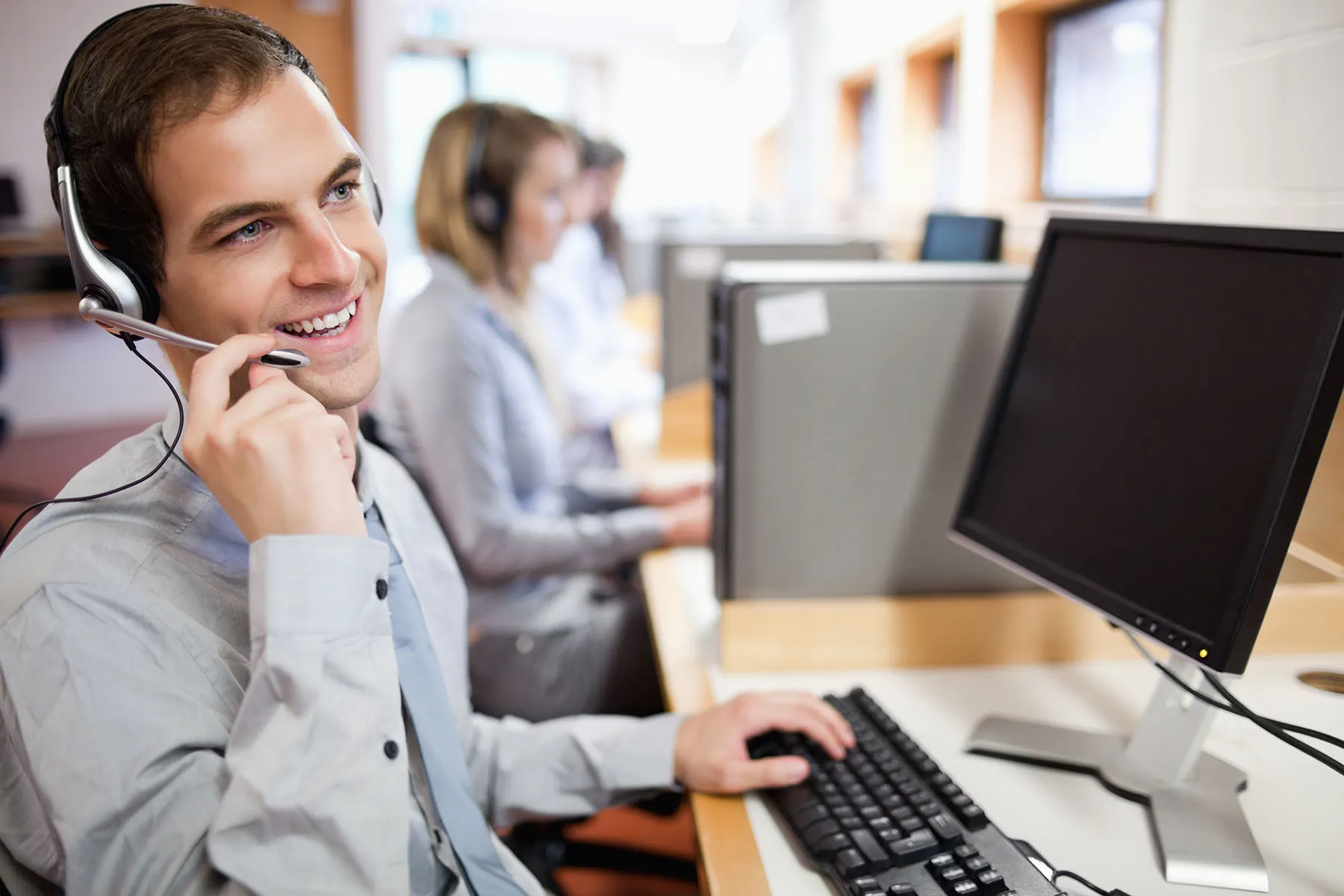 Smiling call center agent wearing a headset at a computer, with other support staff working in the background.