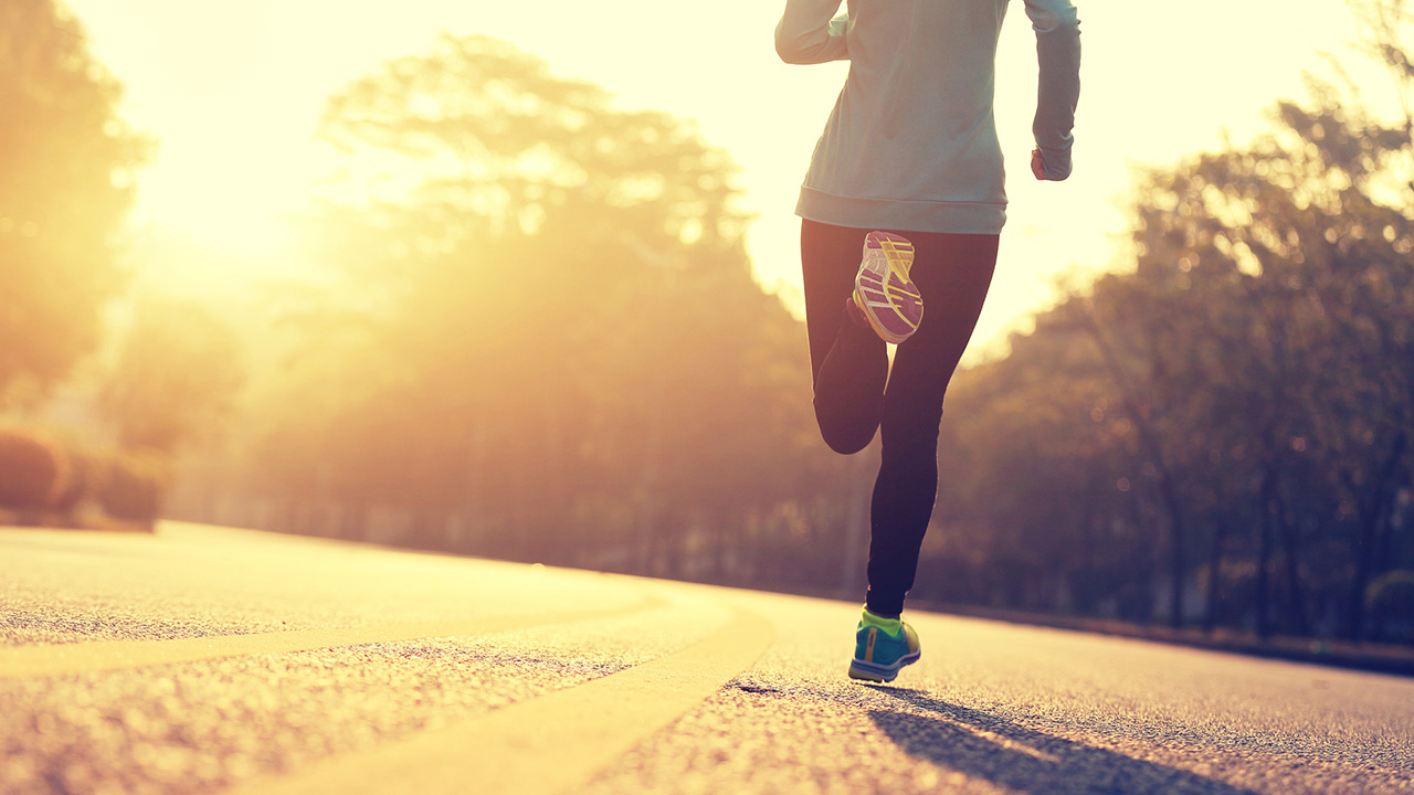 A person is running on a sunlit road, surrounded by blurred trees, under a bright, warm sky.