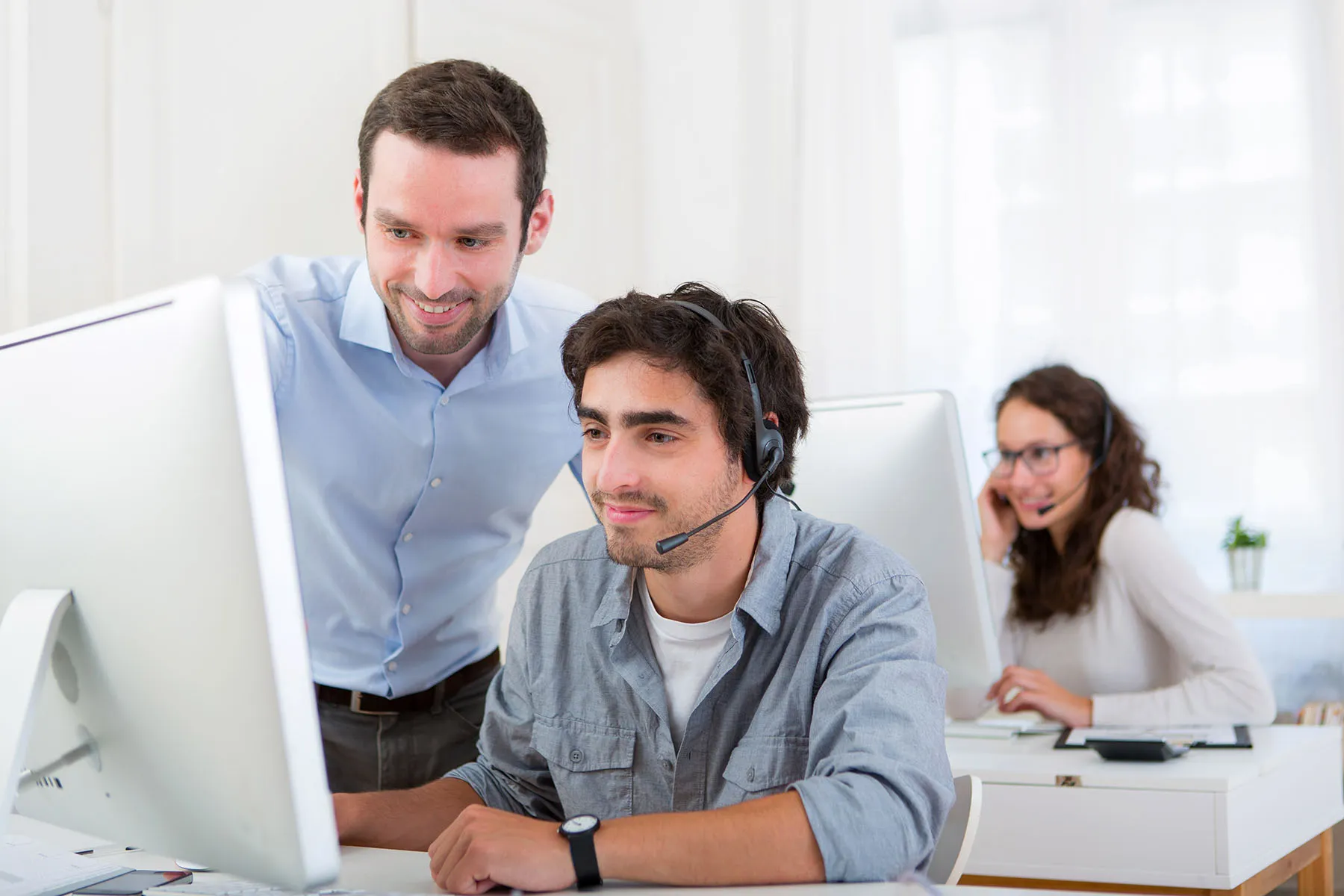 Call center team member wearing a headset at a computer while a supervisor provides support, with another agent in the background.