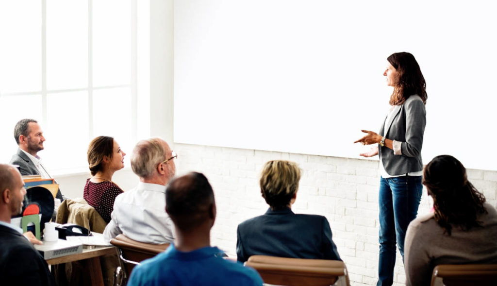 A woman stands and gestures while presenting to a seated group in a well-lit room, with a blank whiteboard behind her.