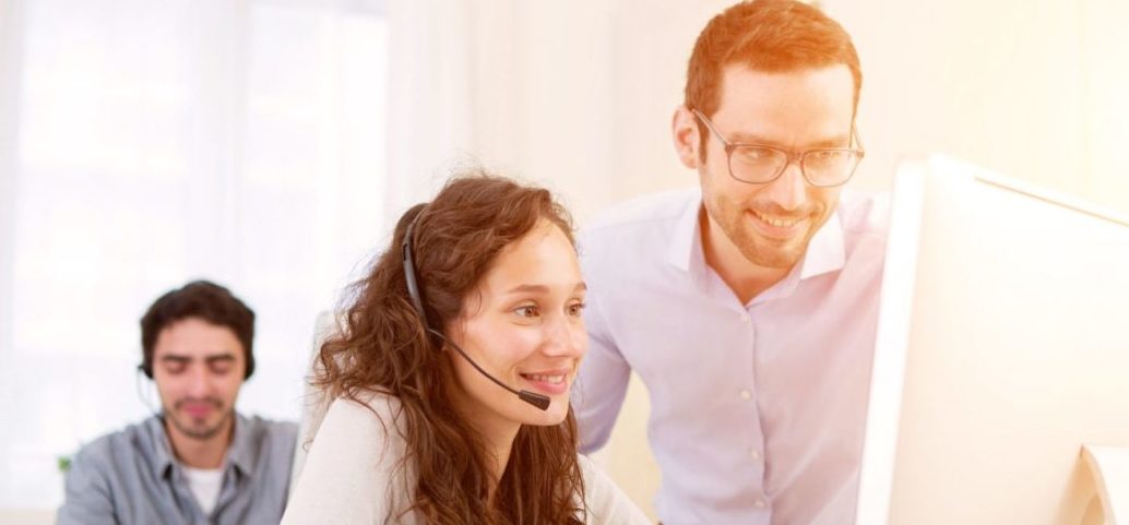 Customer service agent wearing a headset receives guidance from a supervisor while working at a computer, with another agent in the background.