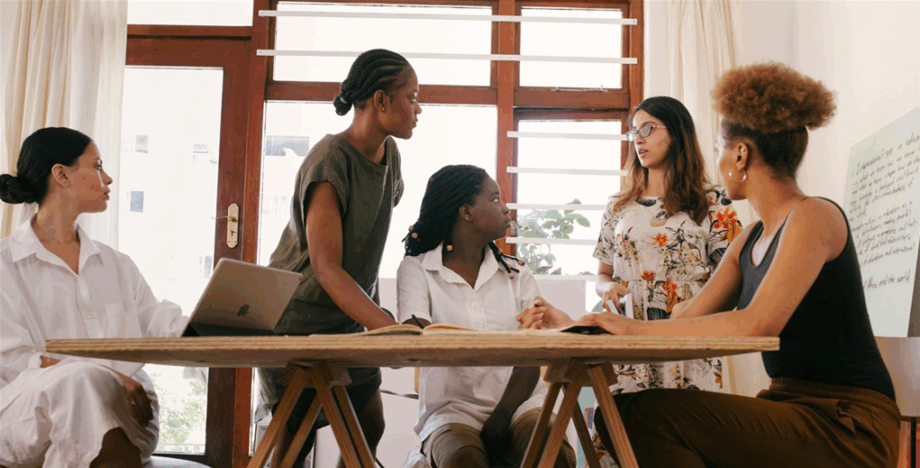 Five people, engaged in discussion around a wooden table, are in a bright room with large windows. One person has a laptop, and a written flipchart is partially visible.