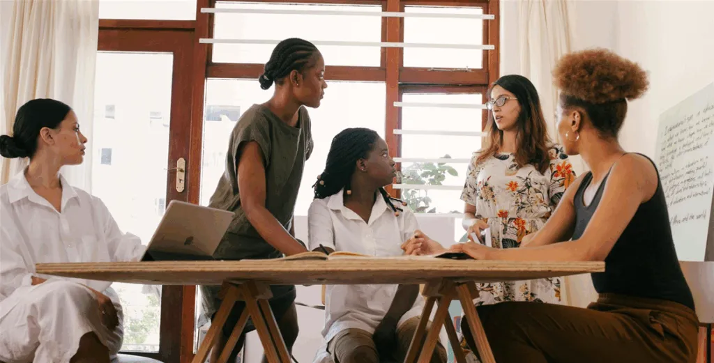 Five people, engaged in discussion around a wooden table, are in a bright room with large windows. One person has a laptop, and a written flipchart is partially visible.