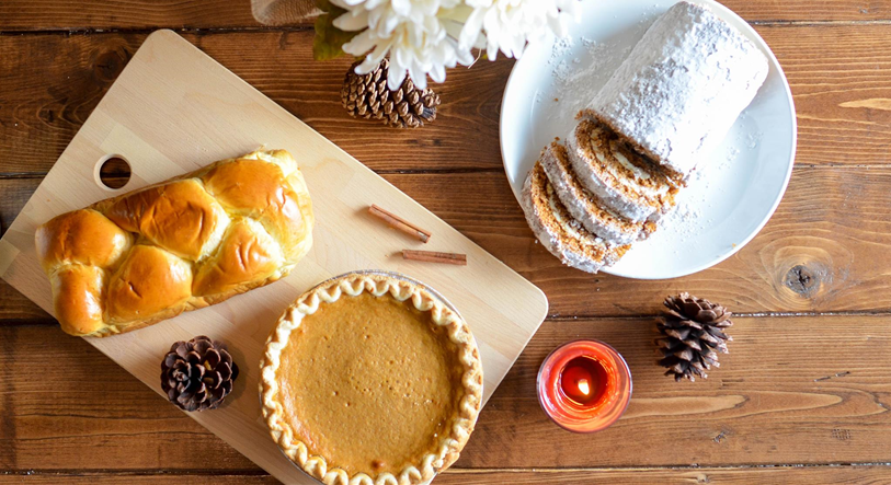 A loaf of braided bread, a pumpkin pie, and a sliced roll dusted with powdered sugar on a wooden table, surrounded by pinecones, cinnamon sticks, and a small red candle.