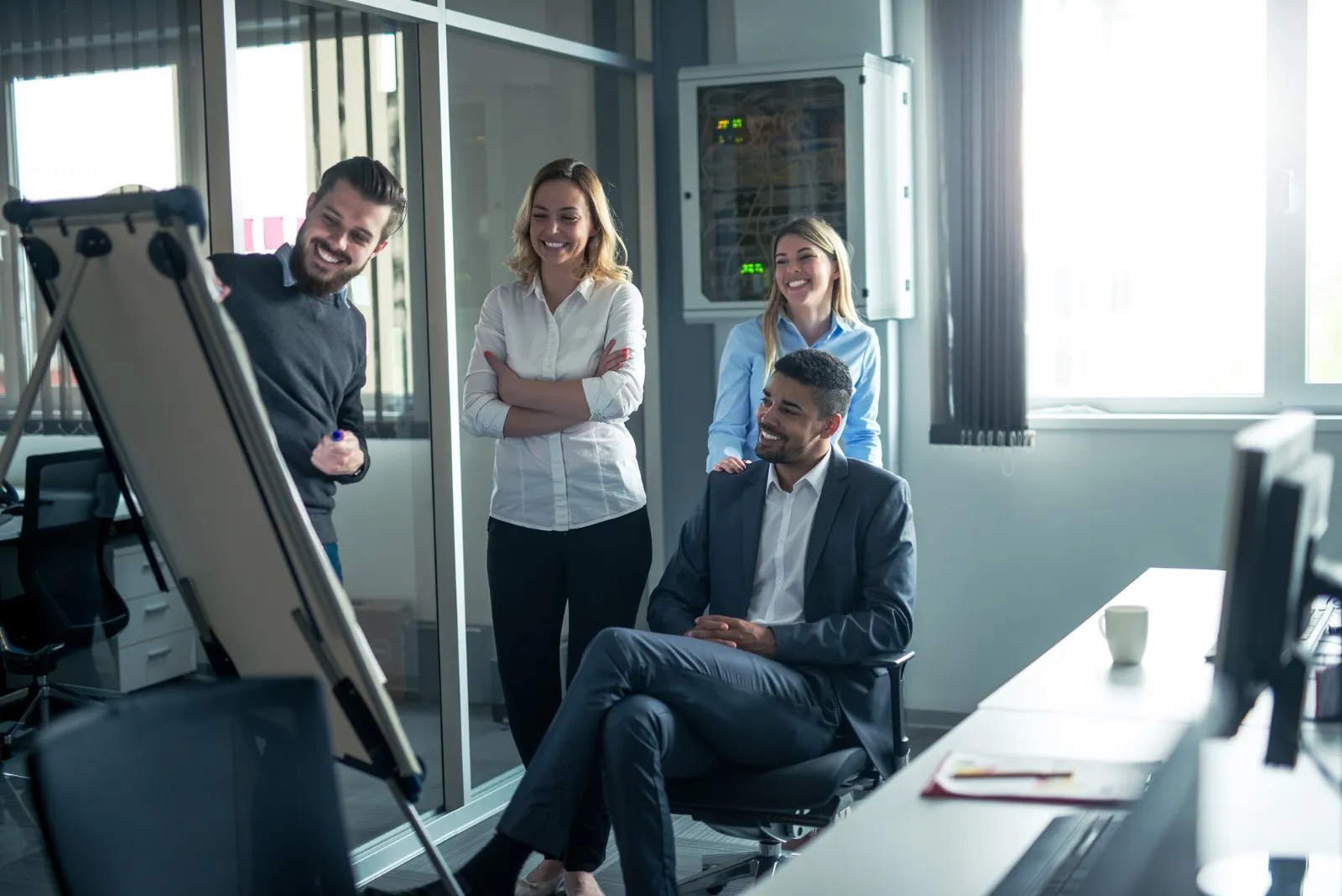 A man presents on a flip chart to three smiling colleagues, one seated, in a modern office with glass partitions and computer monitors.