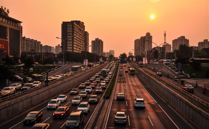 Cars travel along a two-directional highway, surrounded by high-rise buildings at sunset. The sky is a warm orange, creating a contrasting urban evening atmosphere.
