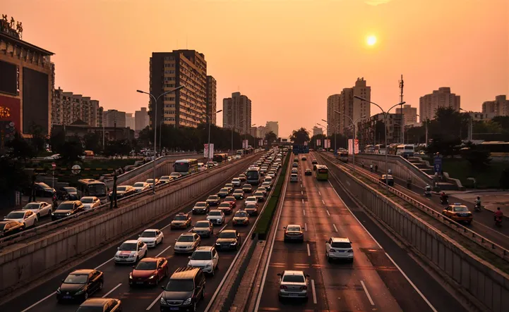 Cars travel along a two-directional highway, surrounded by high-rise buildings at sunset. The sky is a warm orange, creating a contrasting urban evening atmosphere.