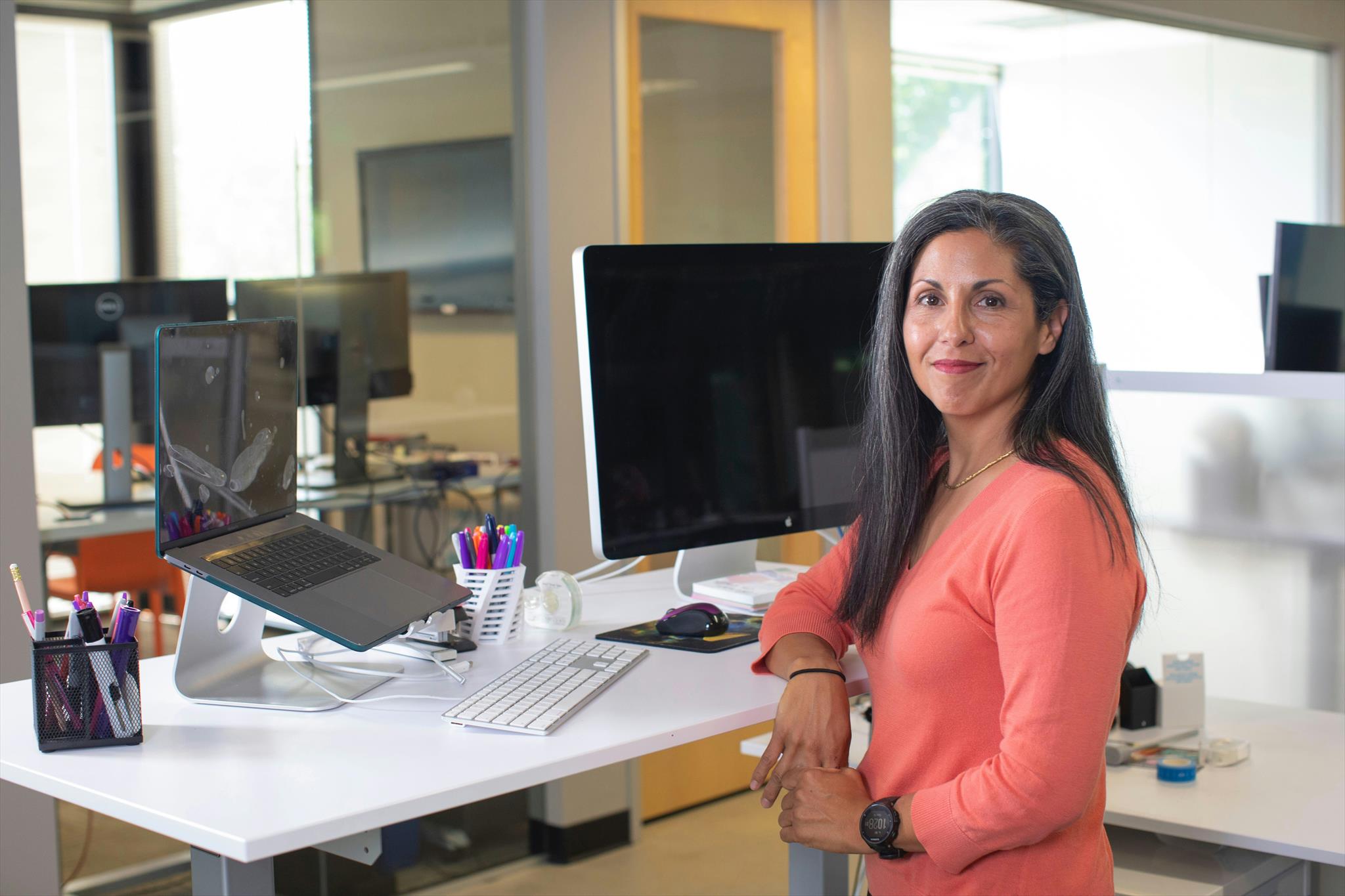 A woman stands confidently at a modern office desk, surrounded by computers, colorful pens, and stationery in a bright, professional workspace.