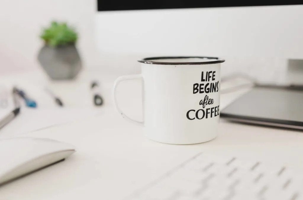 A white mug with the text "LIFE BEGINS after COFFEE" stands on a desk, surrounded by papers, pens, a laptop, and a small plant near a computer monitor.