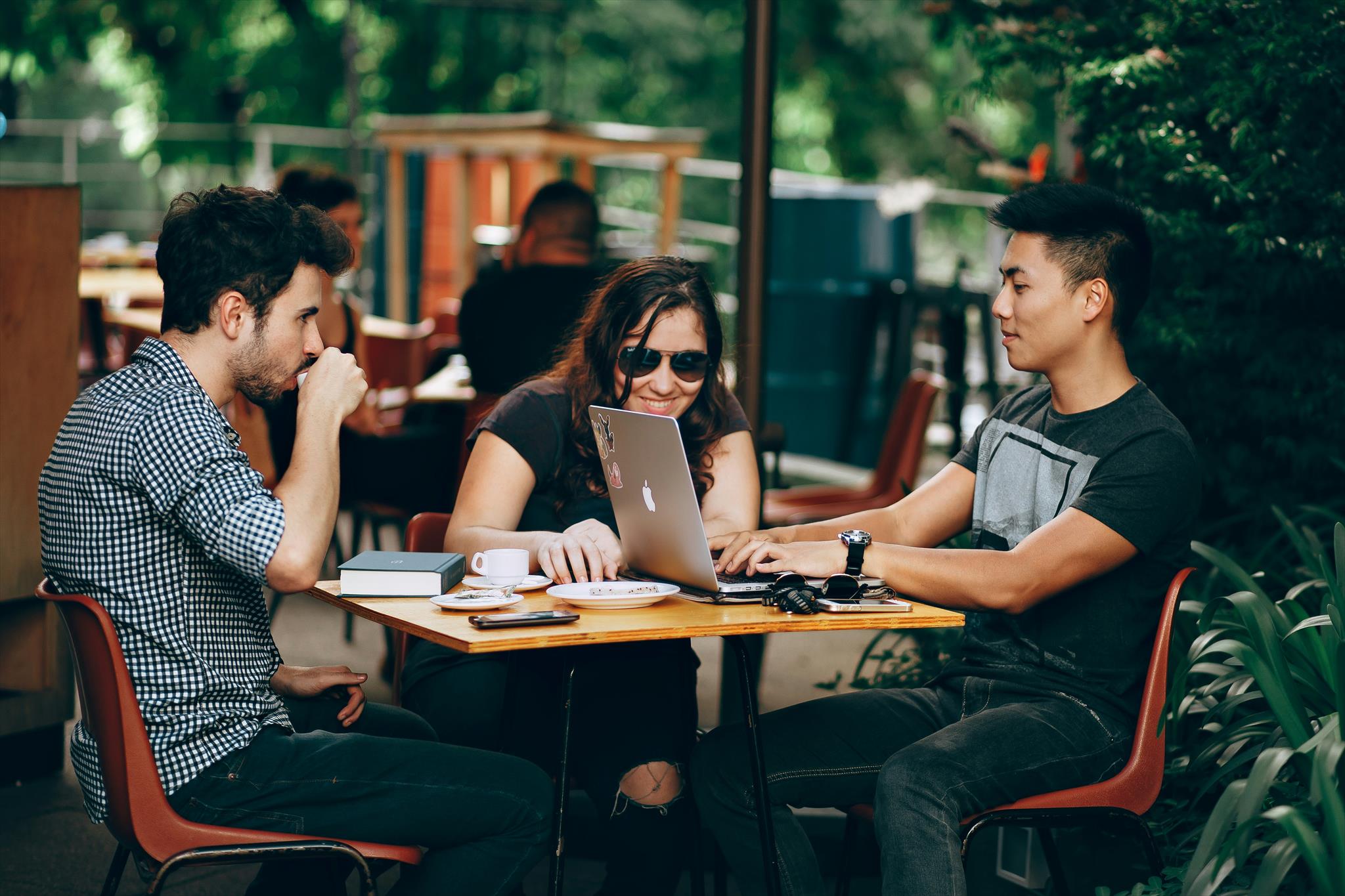 Three people sit at an outdoor table, collaborating on a laptop. Two men and a woman are surrounded by trees and greenery, with coffee cups and notebooks on the table.