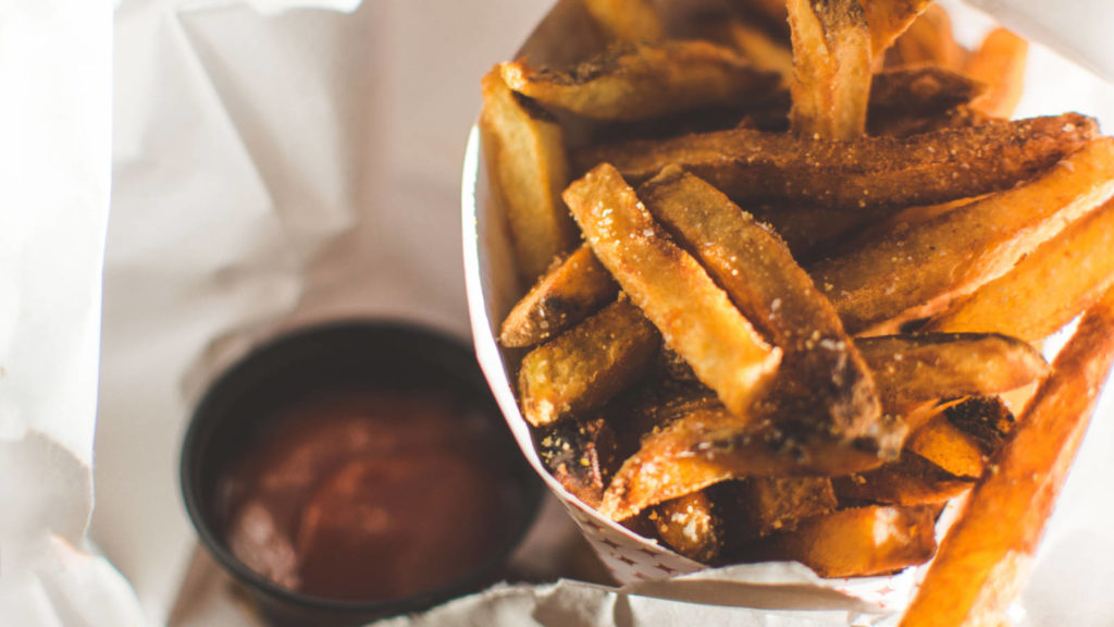 Golden-brown French fries fill a paper cup, accompanied by a small black cup of ketchup, placed on a white, crumpled paper surface.