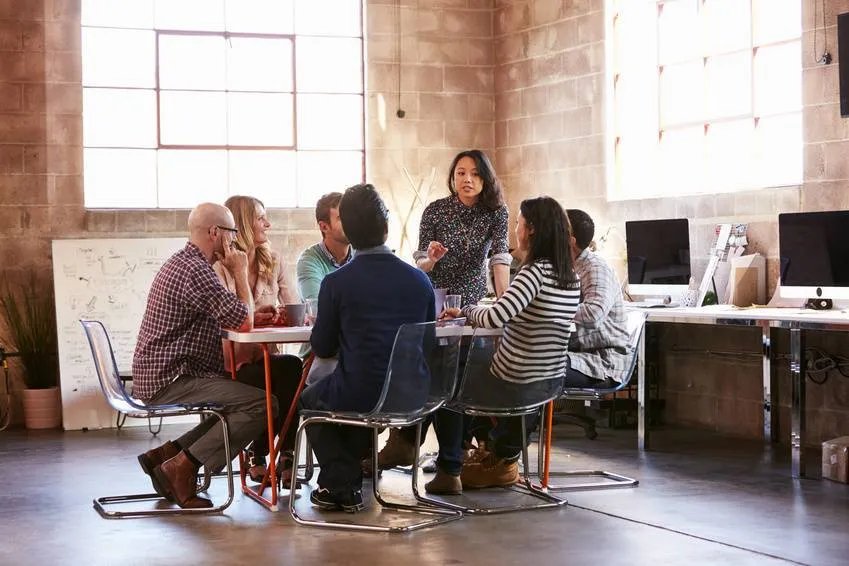 A group of seven people discusses around a table in a modern office with large windows and computers. A whiteboard with diagrams stands nearby.