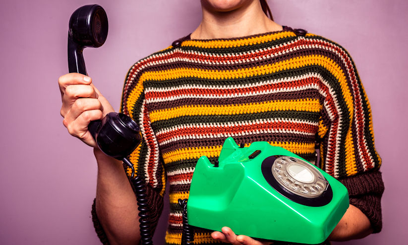A person holding a green rotary telephone, lifting the black receiver, wearing a striped sweater, against a purple background.