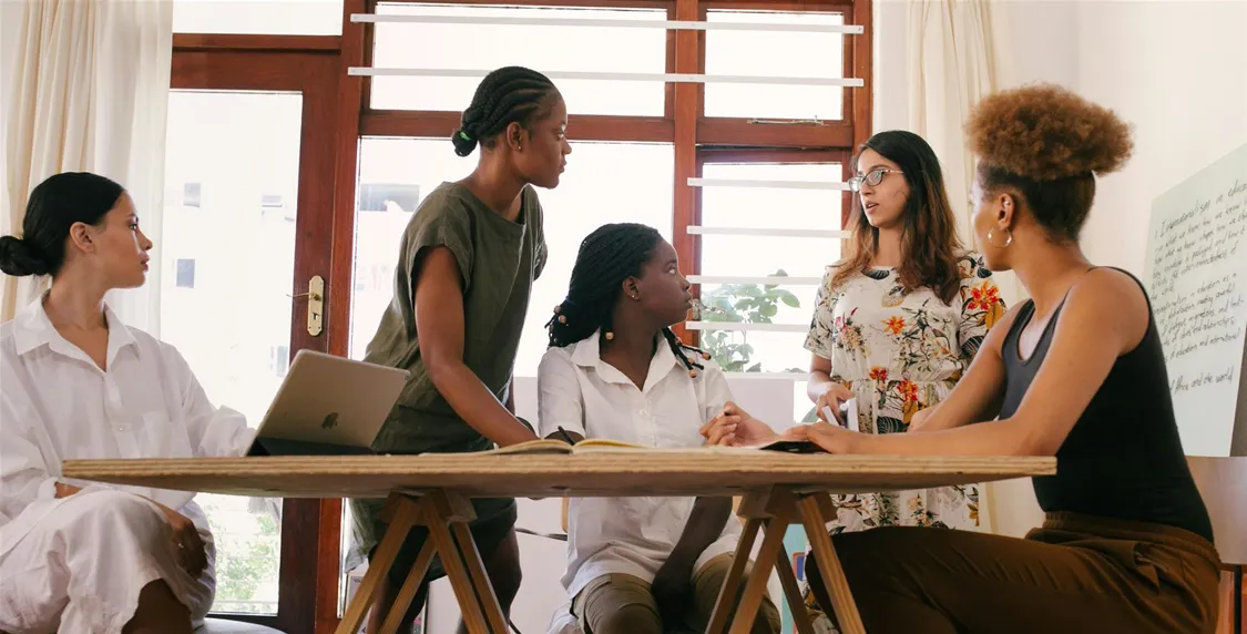 Five people, engaged in discussion around a wooden table, are in a bright room with large windows. One person has a laptop, and a written flipchart is partially visible.